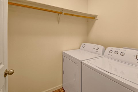 Small laundry room here at Phoenix Ridge Apartment Homes with white washer and dryer against a beige wall. A wooden shelf with a hanging rod is above the appliances. The space is tidy and functional.