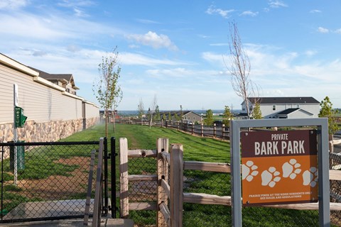 A fenced dog park here at Prairie Pines Apartment Homes with a "Private Bark Park" sign features paw prints. The area has green grass, a few young trees, and is set against a clear blue sky.
