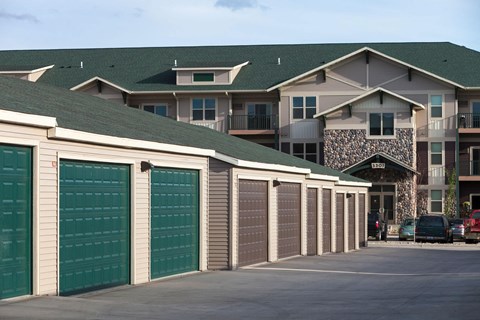 Prairie Pines Apartment Homes complex with stone facade and green roofs, featuring a row of garages in the foreground. Cars are parked, creating a quiet suburban feel.