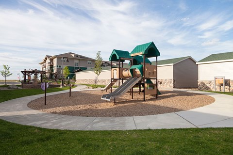 Children's playground here at Prairie Pines Apartment Homes with slides and climbing structures on mulch, surrounded by a path and grass. Modern apartment buildings are visible in the background.