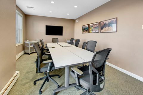 Modern conference room here at Prairie Pines Apartment Homes with beige walls, long white table, and ergonomic chairs. A flat-screen TV and landscape art are mounted on the wall. Cozy and professional ambiance.