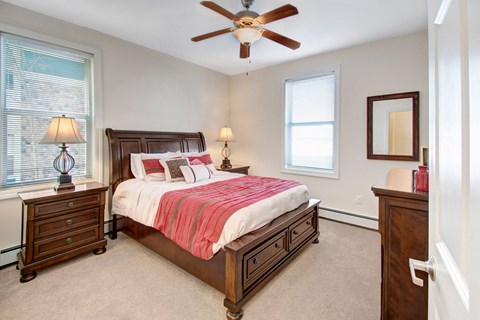 Cozy bedroom here at Prairie Pines Apartment Homes with a wooden bed adorned with red and white bedding. Two matching nightstands with lamps, a ceiling fan, and two windows create a warm ambiance.