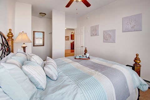 Cozy bedroom here at Prairie Pines Apartment Homes with pastel bedding, wooden bedposts, and a ceiling fan. Wall art features seashells. A tray with a cup sits on the bed, creating a serene vibe.