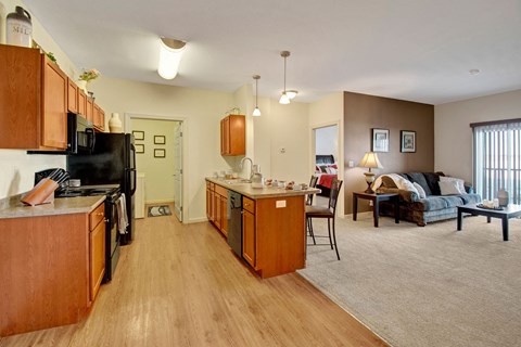 Open-concept living space here at Prairie Pines Apartment Homes with a kitchen featuring wooden cabinets, black appliances, and a central island. Cozy living area with a sofa, lamp, and coffee table, lit warmly.
