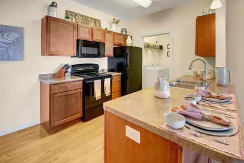 Warm kitchen here at Prairie Pines Apartment Homes with wooden cabinets, black appliances, and a laundry room in the background. A countertop bar features neatly set dishes and decor.