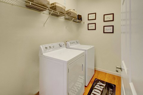 A small, tidy laundry room here at Prairie Pines Apartment Homes with a washer and dryer side by side, a shelf above holding baskets, and four framed quotes on the wall. Warm, organized atmosphere.