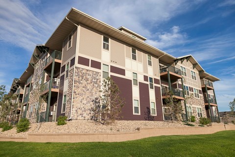 Modern three-story Prairie Pines Apartment Homes building with stone and wood paneling, surrounded by landscaped greenery under a partially cloudy blue sky.