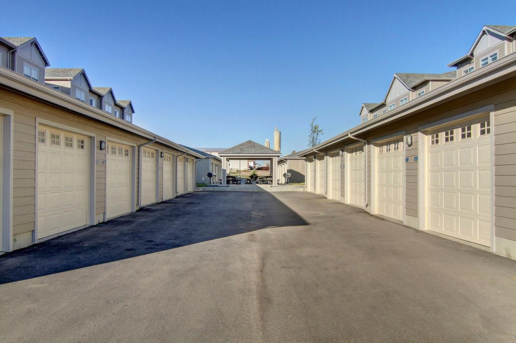 A symmetrical row of beige garages here at Prairie Vista Apartment Homes lines both sides of an asphalt driveway, leading to a small pavilion with picnic tables under a clear blue sky.