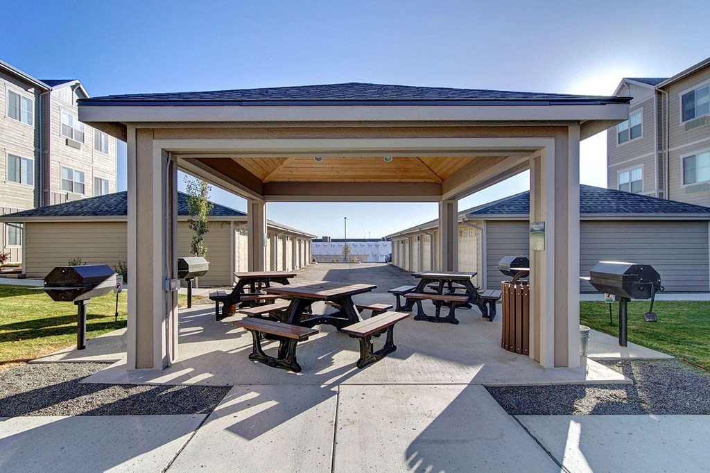 Covered outdoor area here at Prairie Vista Apartment Homes with picnic tables and grills surrounded by residential buildings. The scene is bright and calm, suggesting a communal space for gatherings.