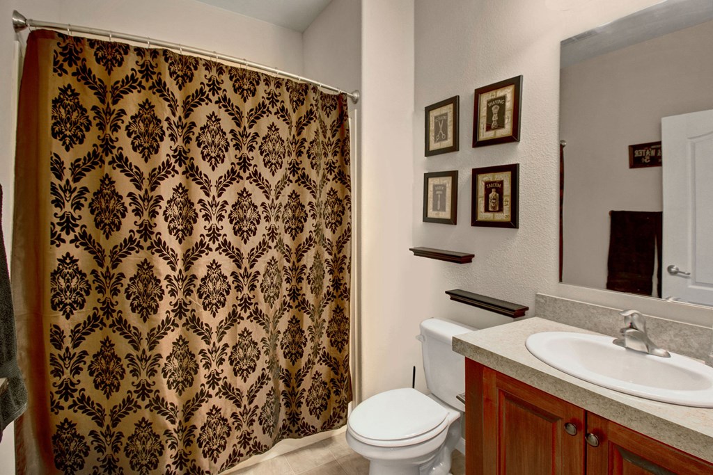 Bathroom here at Prairie Vista Apartment Homes with a beige and black floral shower curtain, white countertop with sink, wooden cabinets, and framed pictures, conveying elegance and warmth.