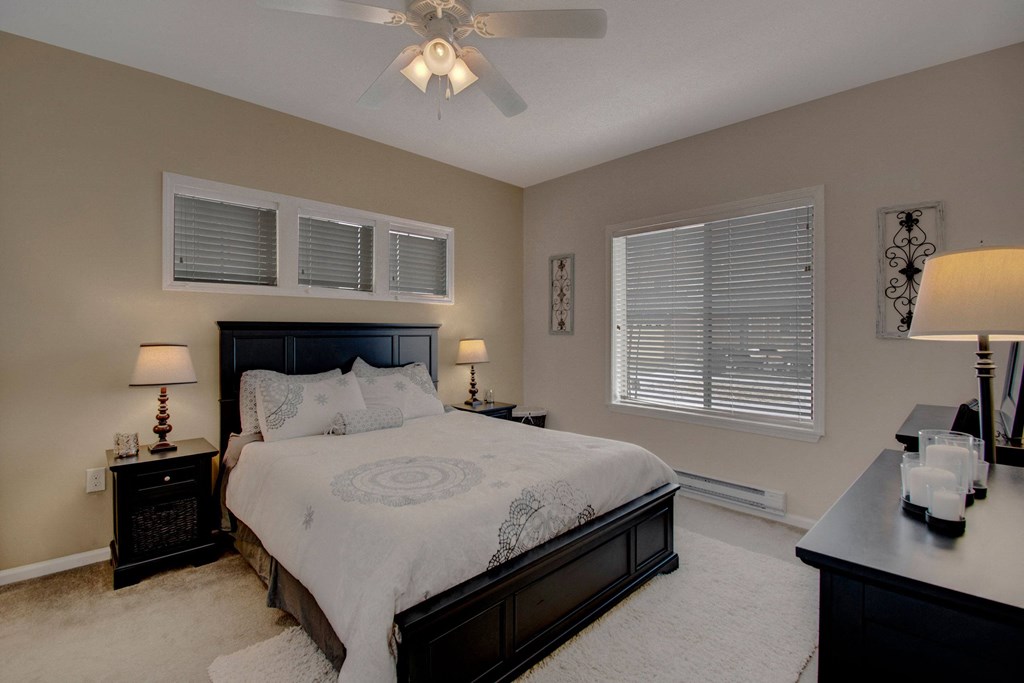 Cozy bedroom here at Prairie Vista Apartment Homes with beige walls, featuring a plush bed with patterned white linens. Black furniture accents, soft lamp lighting, and window blinds add warmth.