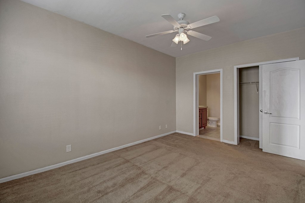 Empty bedroom here at Prairie Vista Apartment Homes with beige carpet and walls, a ceiling fan with lights, open closet with a white door, and a partial view of a bathroom. Calm and neutral tone.