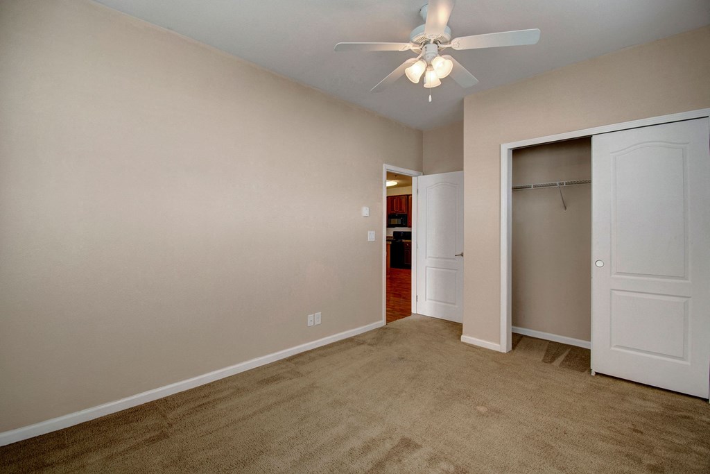 Empty beige bedroom here at Prairie Vista Apartment Homes with tan carpet, white ceiling fan, and open closet. A doorway reveals a glimpse of a kitchen with dark cabinets. Calm ambiance.