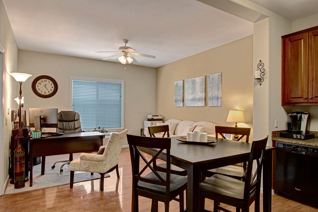 A cozy home office and dining area here at Prairie Vista Apartment Homes. A desk with a computer and chair is by the window, with a clock on the wall. A dining table set is nearby, and soft lighting creates a welcoming ambiance.