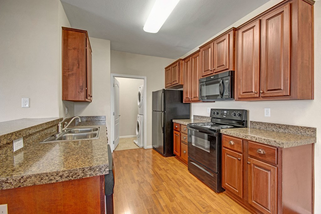 Modern kitchen here at Prairie Vista Apartment Homes with dark wood cabinets, granite countertops, and black appliances, featuring a stove, microwave, and fridge. A stackable washer-dryer is visible in an adjacent room.