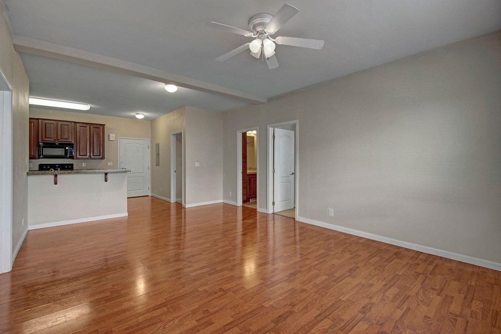 Spacious empty room here at Prairie Vista Apartment Homes with wooden floors and neutral walls. Ceiling fan overhead. Open plan leads to kitchen with dark cabinets and white counter.