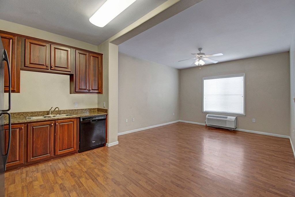 Spacious room here at Prairie Vista Apartment Homes with wooden floors and neutral walls, featuring a corner kitchen with dark wood cabinets, granite countertops, and a ceiling fan above.