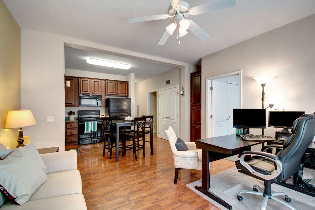 A cozy living space here at Prairie Vista Apartment Homes with wood flooring features a sofa, a desk with dual monitors, and a comfortable chair. In the background, there's a well-lit kitchen with wooden cabinets and a dining table. The room has a warm, inviting ambiance with modern decor and ceiling fan.
