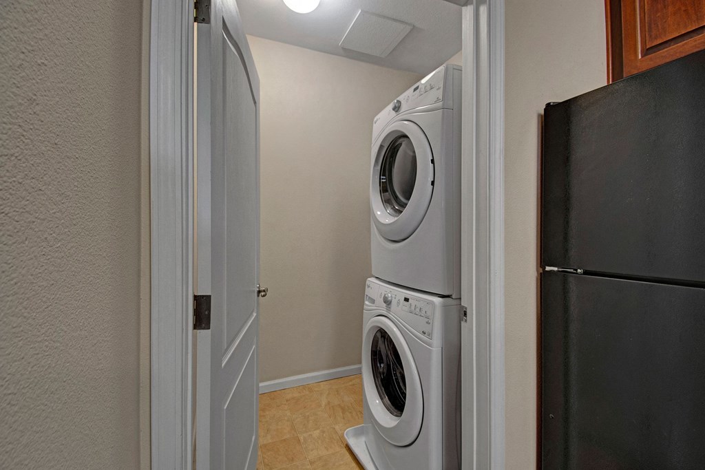 Stacked washer and dryer in a small laundry nook here at Prairie Vista Apartment Homes with beige walls and tile flooring. A black refrigerator is partially visible on the right.