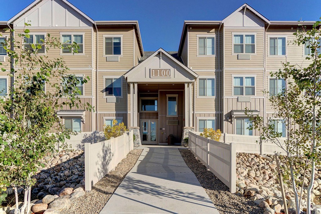 A three-story, tan Prairie Vista Apartment Homes building with a central entrance labeled "1100" sits under a clear blue sky. A pathway flanked by white fences and rocks leads to the door, with small trees on either side.