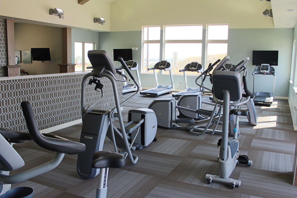 Bright gym room here at Renaissance Heights Apartment Homes with large windows showing an outdoor view. Equipped with treadmills, ellipticals, and exercise bikes. Modern, serene atmosphere.
