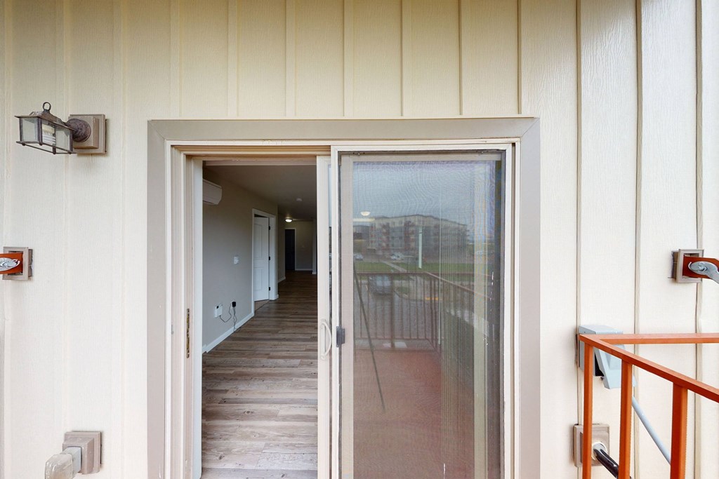 View from a balcony through an open sliding door here at Renaissance Heights Apartment Homes, revealing a hallway with wooden floors and neutral walls. A cozy, inviting atmosphere is conveyed.