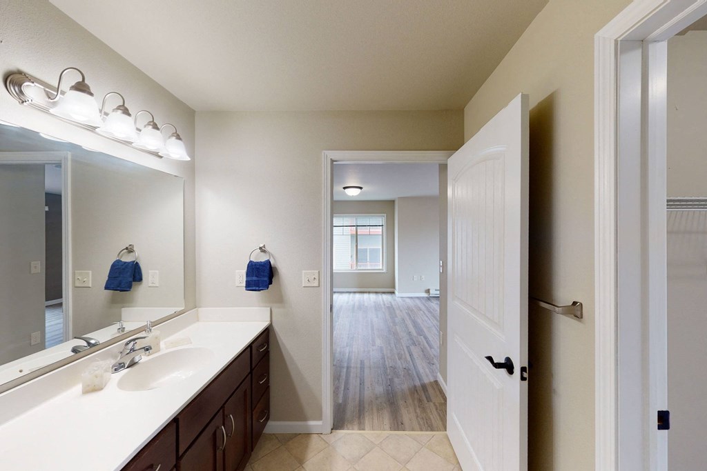 Modern bathroom here at Renaissance Heights Apartment Homes with a large mirror and dual sinks, leading to a bright room with wooden flooring. Blue towels and warm lighting create a cozy feel.