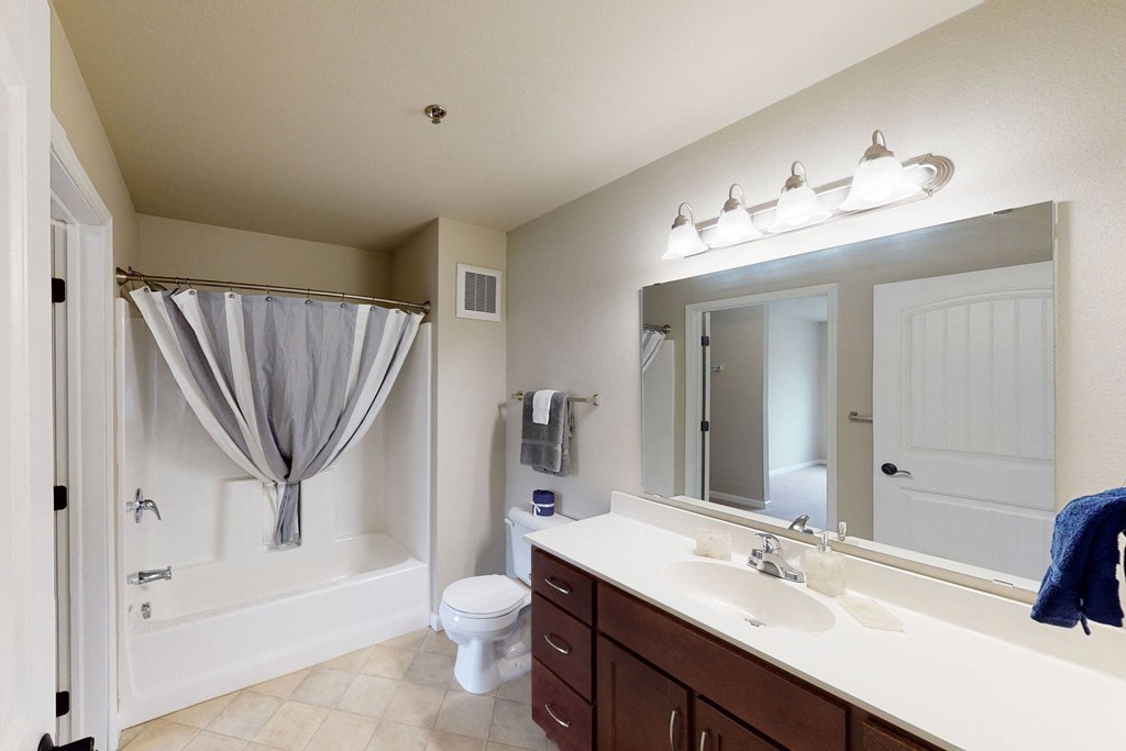 Simple and clean bathroom here at Renaissance Heights Apartment Homes with a bathtub and striped curtain, wooden vanity with double sinks, large mirror, and bright lighting, conveying a fresh feel.
