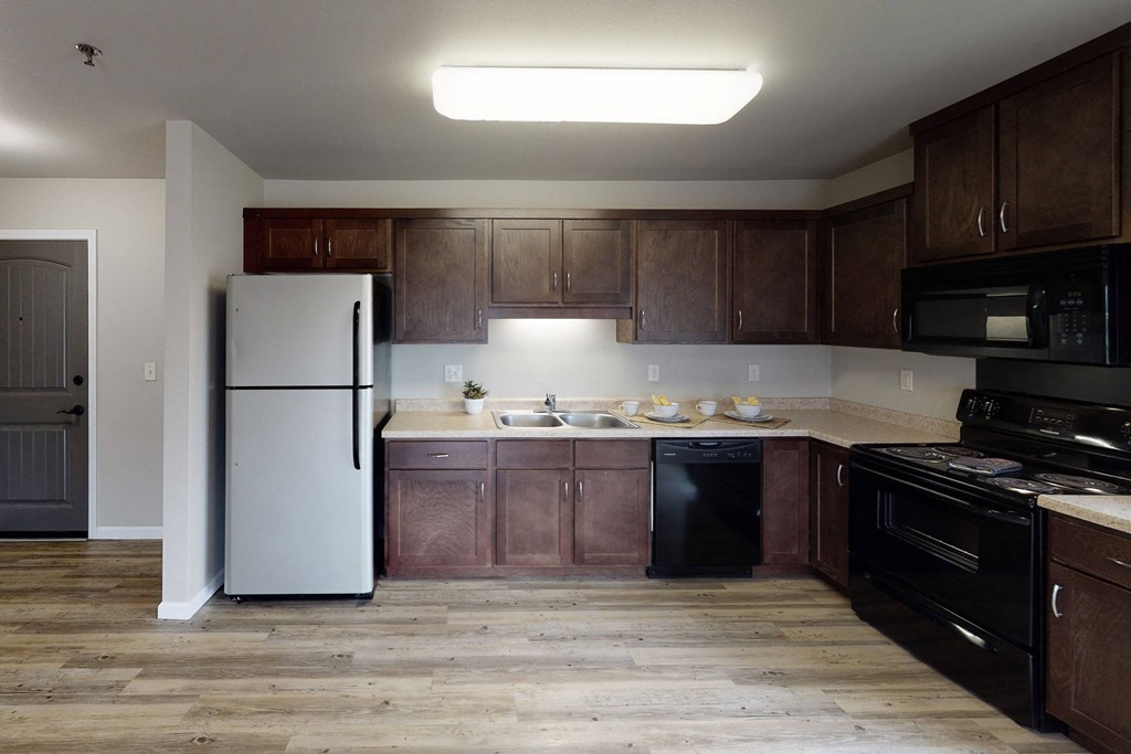 Kitchen here at Renaissance Heights Apartment Homes with wooden cabinets and flooring, stainless steel fridge, black stove and dishwasher, and a sink. Bright, clean, and modern ambiance.
