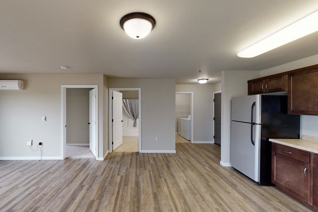 Modern apartment interior here at Renaissance Heights Apartment Homes with wooden flooring and off-white walls. Features include an air conditioner, open kitchen with stainless steel fridge, and a view into a laundry area. The overall tone is clean and inviting.