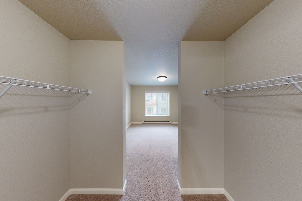 A narrow walk-in closet here at Renaissance Heights Apartment Homes with beige walls and carpet, featuring white wire shelves on both sides, opens to a bright room with a window and overhead light.