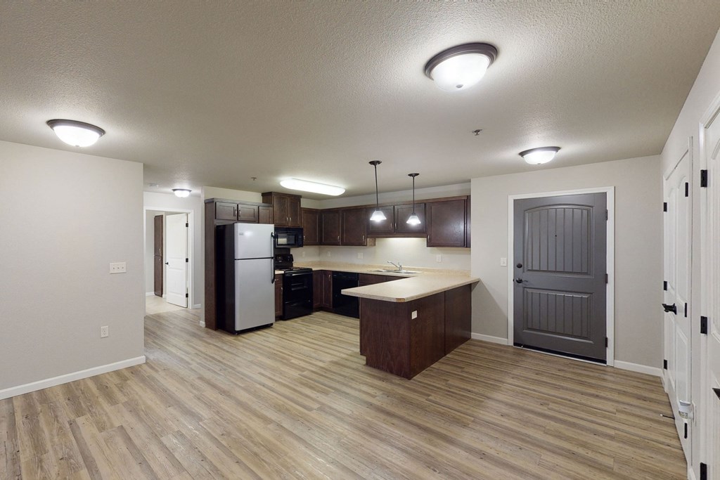 Modern open-plan kitchen here at Renaissance Heights Apartment Homes with wood floors, dark cabinets, stainless steel appliances, and pendant lights over a countertop. Neutral walls, cozy atmosphere.