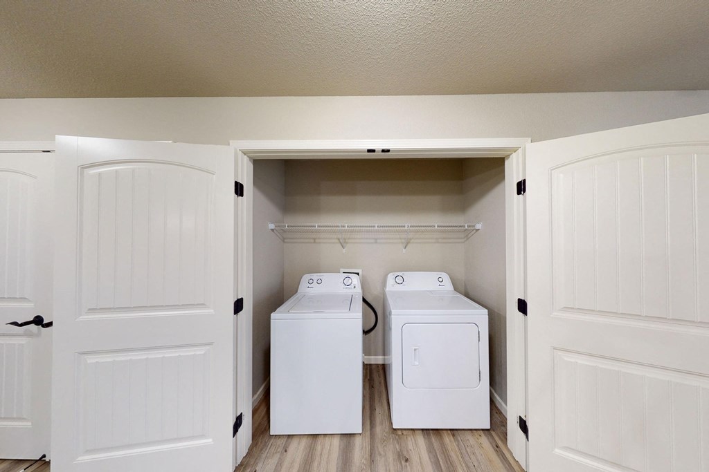 Laundry closet here at Renaissance Heights Apartment Homes with open double doors revealing a white washing machine and dryer on a wooden floor. A single shelf is mounted above.