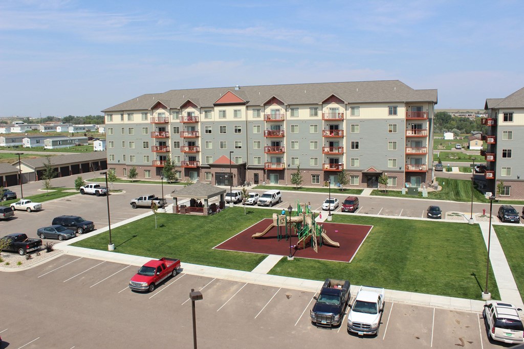 Renaissance Heights Apartment Homes complex with four stories, featuring balconies and a central playground. Cars are parked in adjacent lots under a clear blue sky.