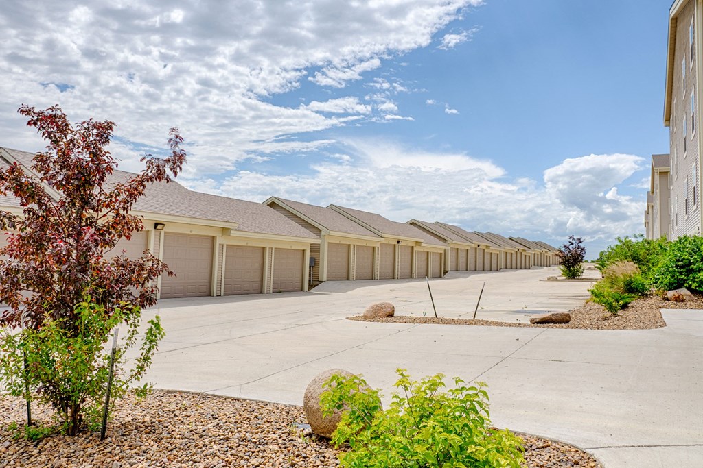Row of beige garage units and a multi-story building here at Rockridge Apartment Homes on a sunny day, with a clear blue sky and scattered clouds. Landscaping includes rocks, small trees, and shrubs.