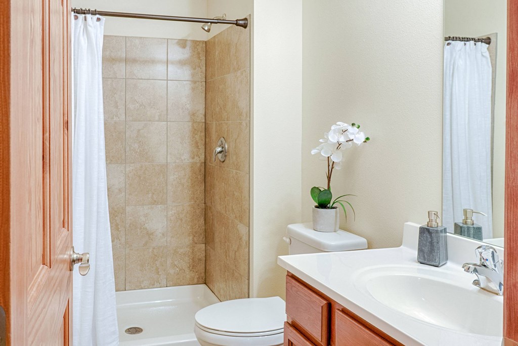 Compact bathroom here at Rockridge Apartment Homes with beige tile shower, white curtain, and wooden cabinet. A white orchid sits on the toilet, adding elegance and calm.
