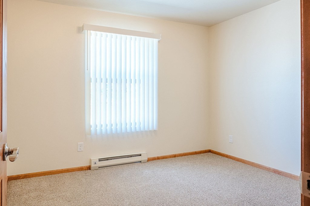Empty bedroom here at Rockridge Apartment Homes with beige walls and carpeted floor, featuring a window with vertical blinds letting in natural light. Simple, neutral, and serene atmosphere.