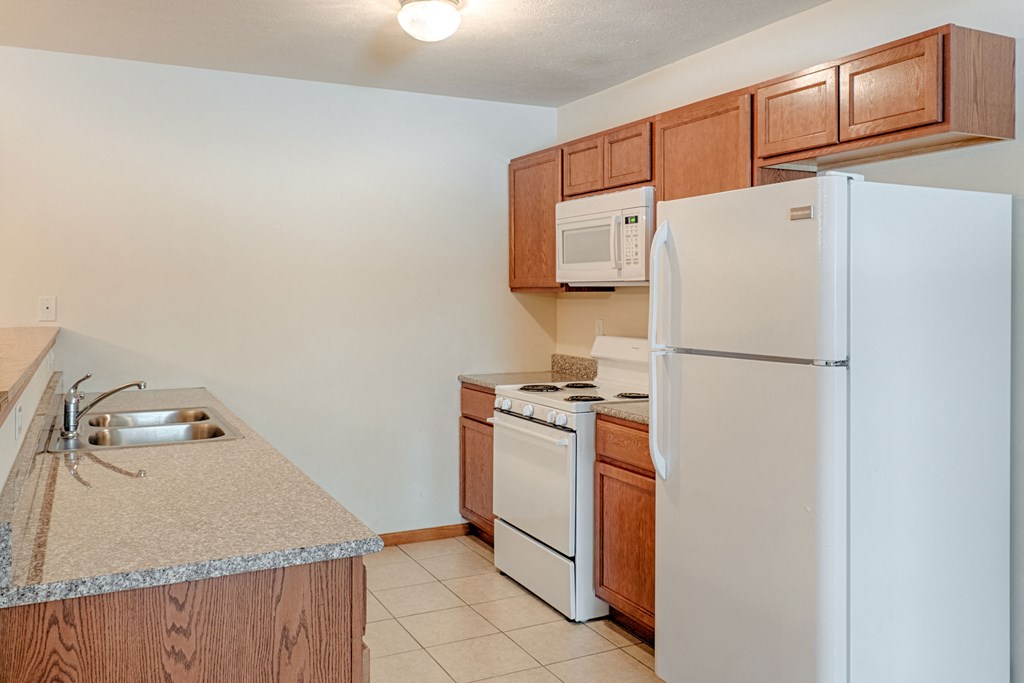 Modern kitchen here at Rockridge Apartment Homes with a white refrigerator, stove, and microwave. Wooden cabinets and marble countertops. Double sink on a tiled floor under soft lighting.