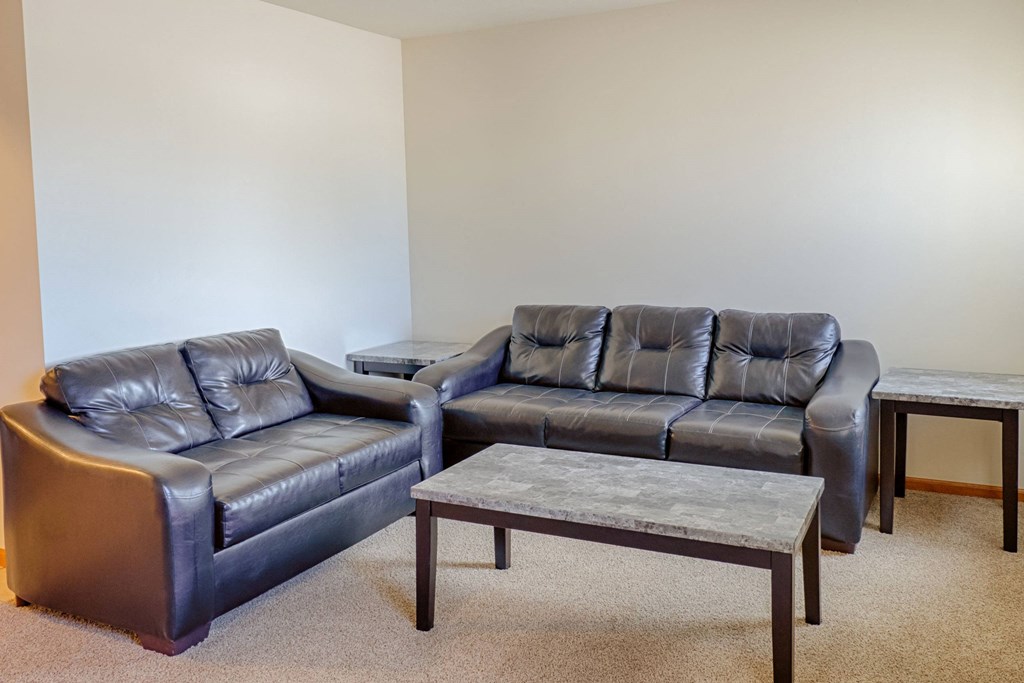 Spacious living room here at Rockridge Apartment Homes with two dark leather sofas, a rectangular coffee table, and two matching side tables on a beige carpet, against a plain wall.
