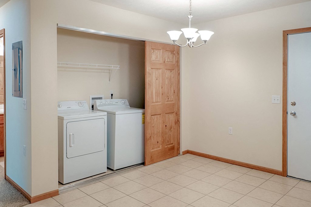 Laundry area here at Rockridge Apartment Homes with a washer and dryer in an open closet, adjacent to a bright room with tiled flooring. A wooden door and modern light fixture are visible.