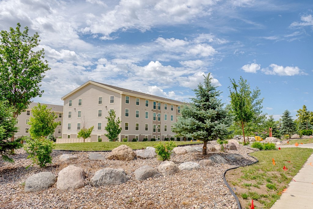 Modern beige Rockridge Apartment Homes building with numerous windows, surrounded by neatly landscaped gardens, rocks, and trees under a bright, partly cloudy sky.