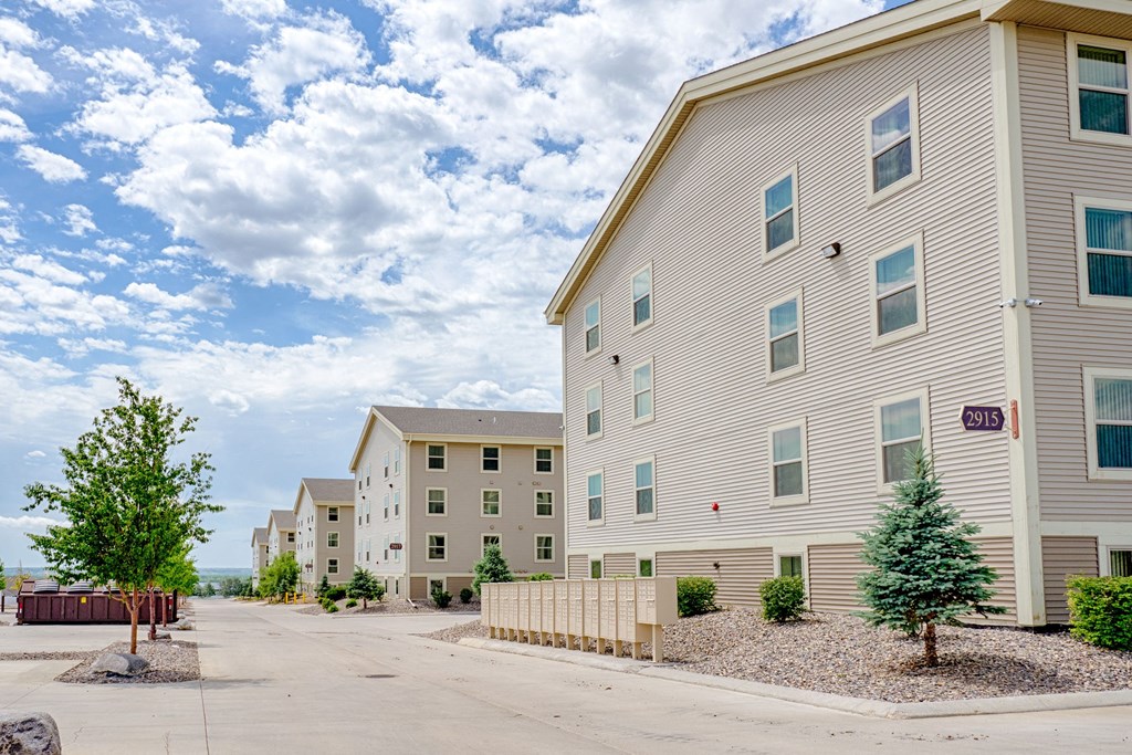 A row of beige Rockridge Apartment Homes buildings lines a quiet street under a partly cloudy sky. Sparse greenery and rocks lie in landscaped areas, conveying a serene, suburban atmosphere.