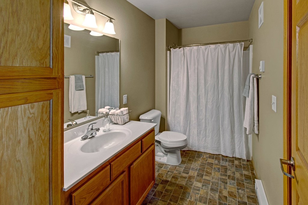 Simple bathroom here at Vue 28 Apartment Homes with warm wood cabinets, white countertop, and sink. A large mirror with lighting, tiled floor, and a white shower curtain create a cozy feel.