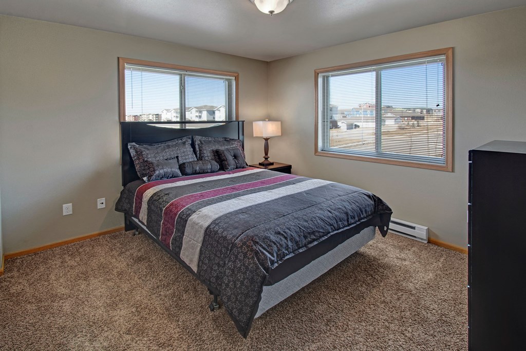 A cozy bedroom here at Vue 28 Apartment Homes with beige walls features a bed with a dark headboard and a striped comforter in shades of gray and burgundy. Two large windows let in ample natural light, and a small lamp on a side table casts a warm glow. The carpet is soft and brown, enhancing the inviting atmosphere.