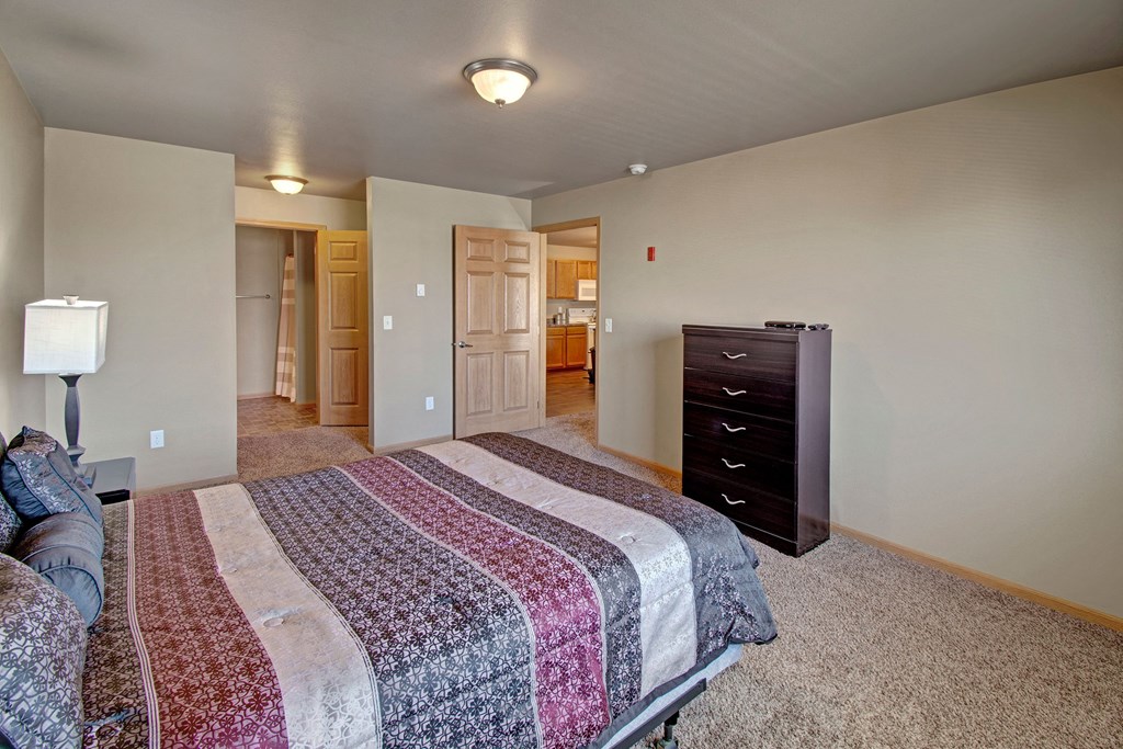 Bedroom here at Vue 28 Apartment Homes with a colorful striped bedspread on a double bed, nightstand with a lamp, and dark chest of drawers. Open door reveals a warm-lit hallway. Neutral, cozy ambiance.
