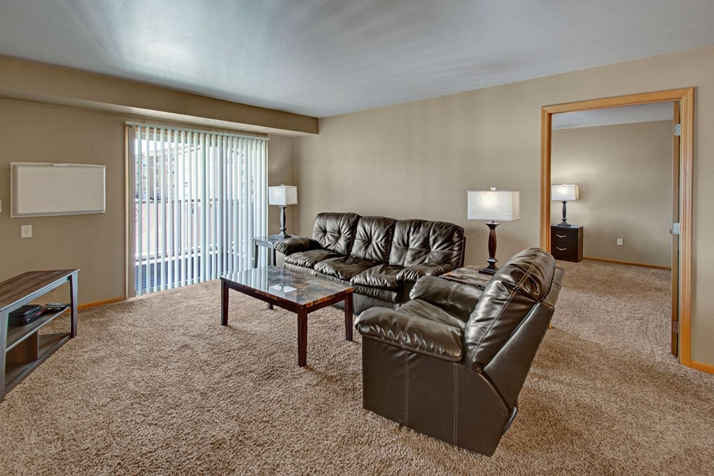 A cozy living room here at Vue 28 Apartment Homes with plush brown leather sofas, a glass coffee table, warm lighting, and beige walls. Vertical blinds cover a window, adding an inviting feel.