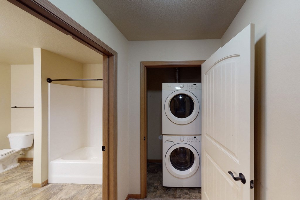 Hallway here at Williston Garden Apartment Homes with a white stacked washer and dryer in an open closet. Nearby, a bathroom with a bathtub, shower, and toilet. Neutral tones create a clean, functional atmosphere.