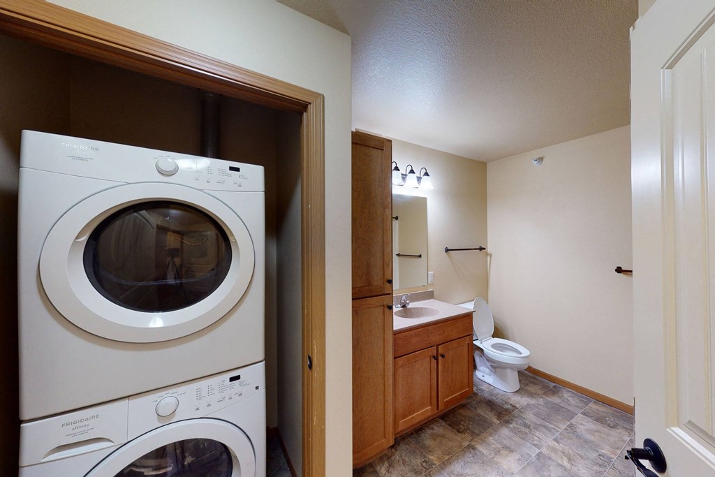 Stacked washer and dryer beside a bathroom with a wooden vanity, mirror, and toilet here at Williston Garden Apartment Homes. The room has a clean, modern look with neutral tones.
