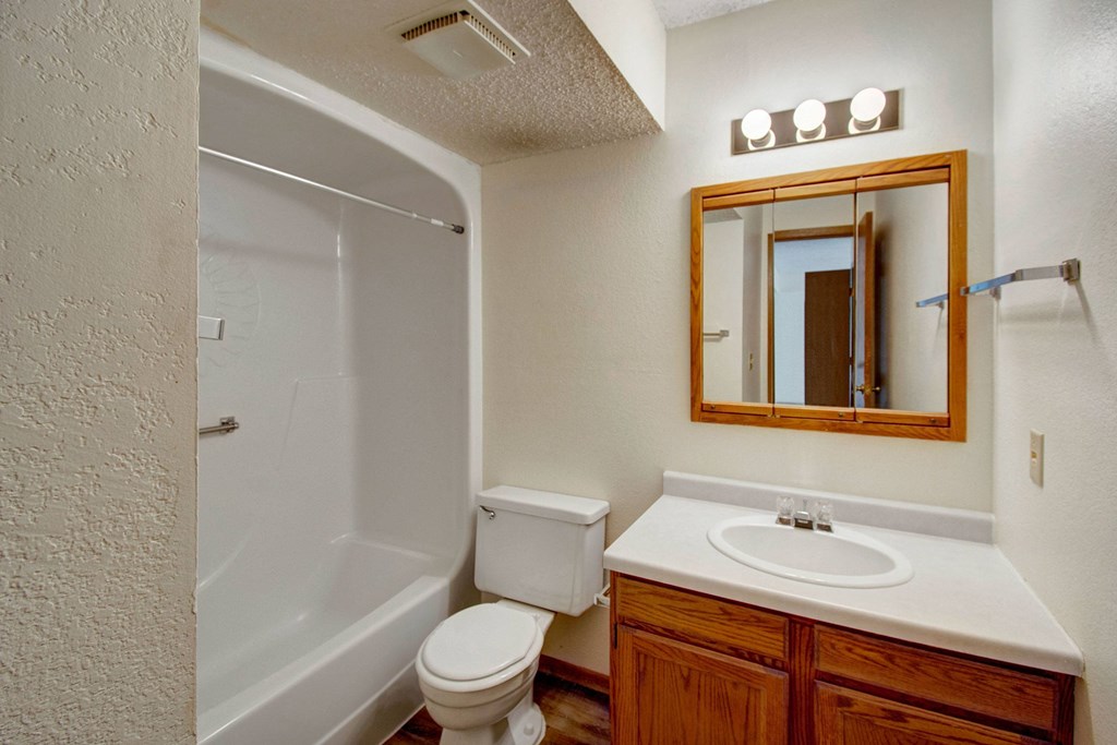 A small, clean bathroom here at Windscape Apartment Homes with a beige color scheme. It features a bathtub with a shower curtain, a toilet, and a wooden vanity with a sink. A mirror cabinet with lights is above the sink, creating a bright and inviting atmosphere.