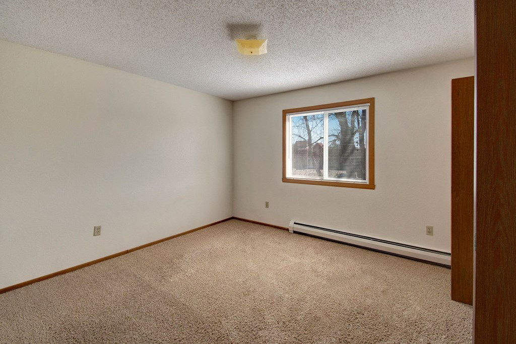 Empty bedroom here at Windscape Apartment Homes with cream walls and beige carpet. A window with wooden trim shows trees and blue sky outside. The space feels calm and minimalistic.